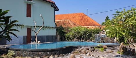Una piscina con borde de piedra, un edificio blanco de techo rojo y follaje circundante.