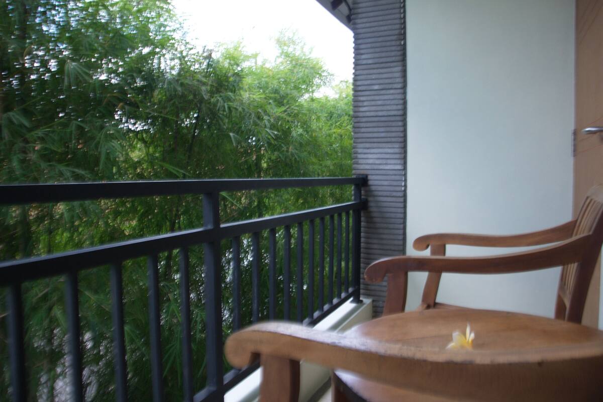 A wooden chair on a balcony with a black railing, overlooking dense green foliage.