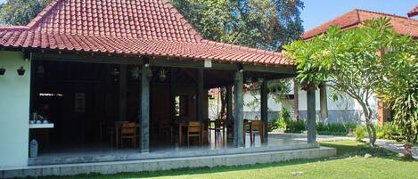 A covered outdoor seating area with tables and chairs, a tiled roof, and a grassy lawn.