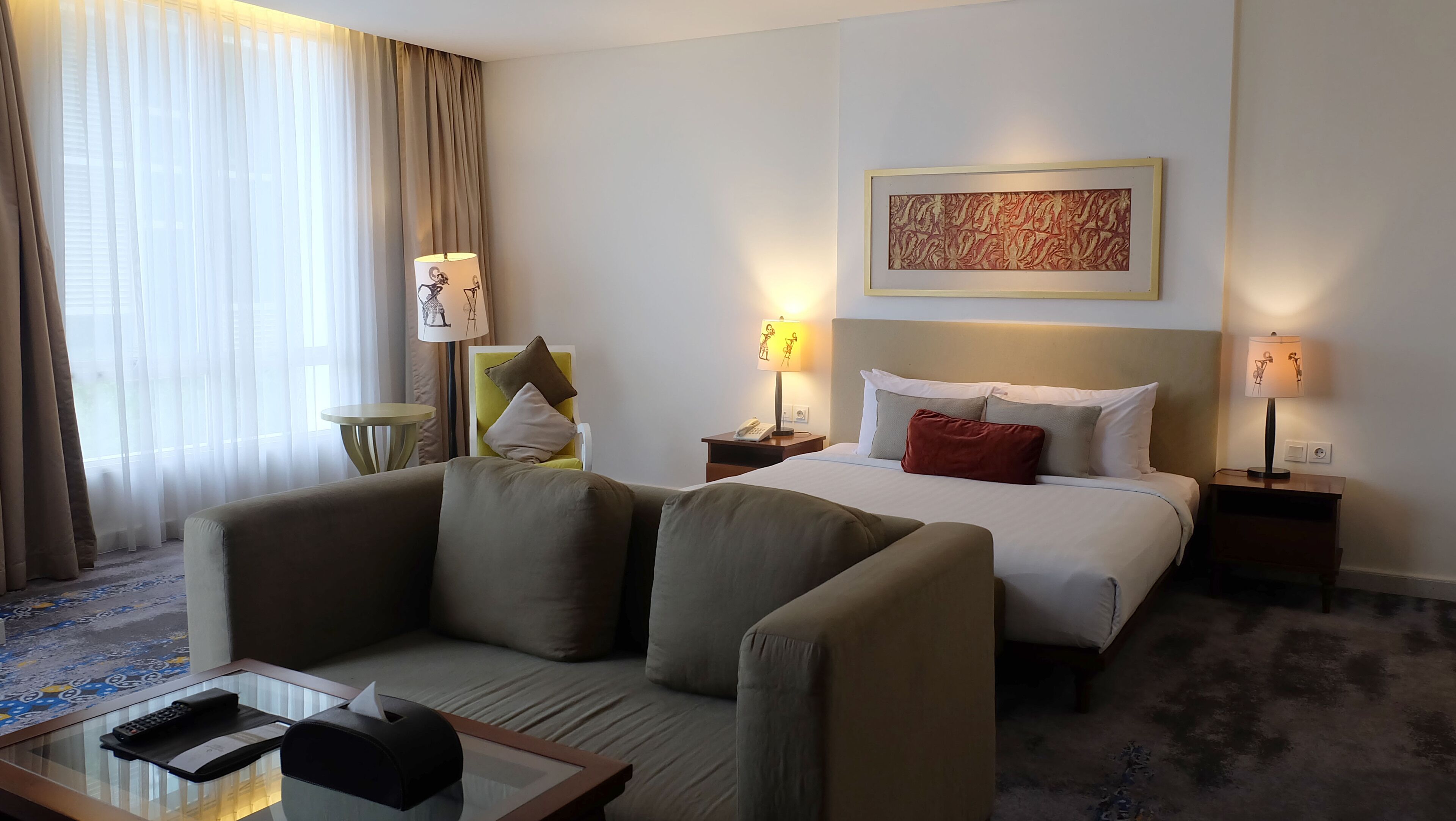A hotel room with a bed, two bedside tables, a lamp, a chair, a small table with a remote control, and a patterned rug.