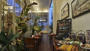 A restaurant with wooden tables and chairs, a variety of dishes displayed on the counter, and a decorative plant arrangement.
