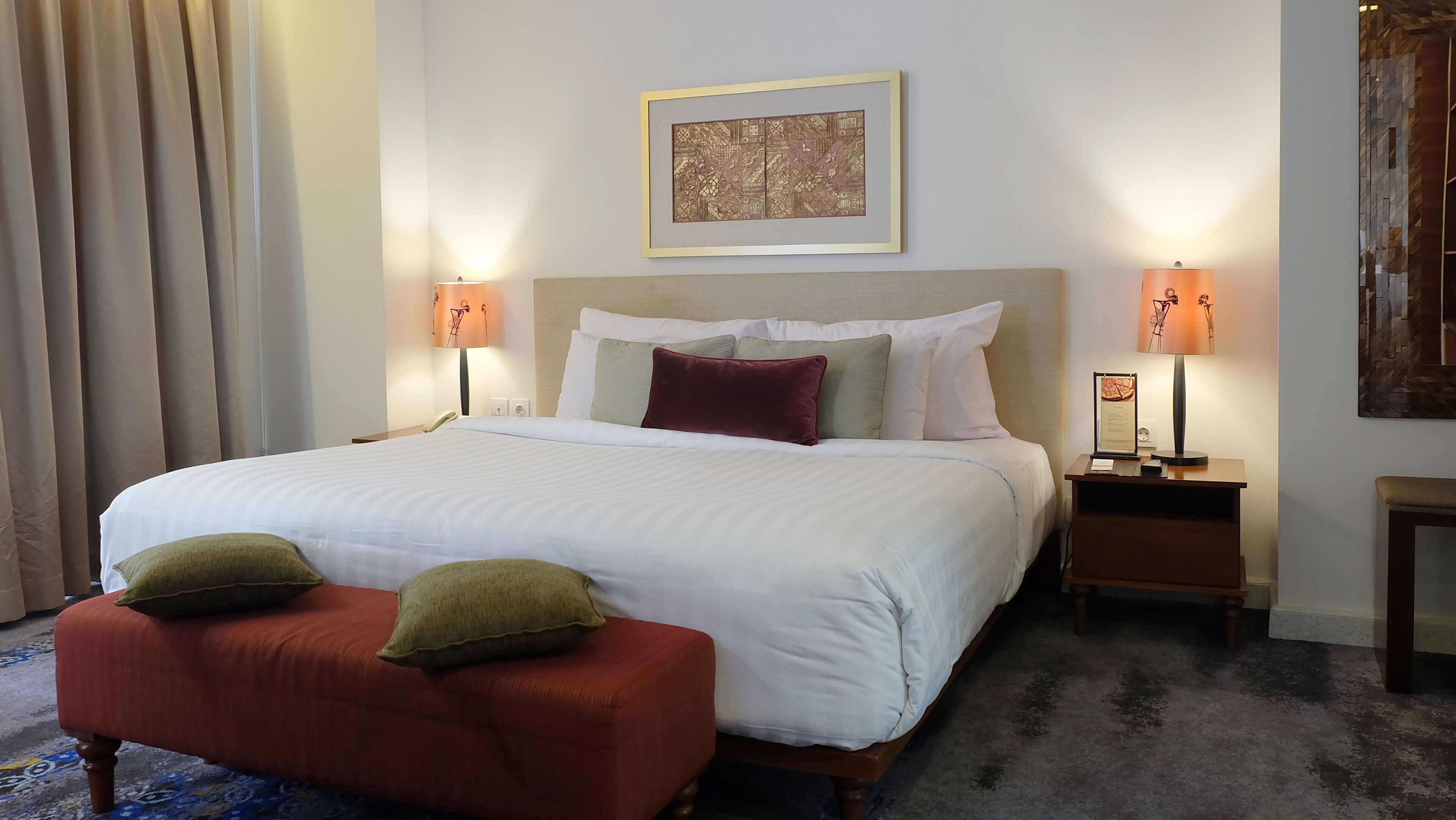 Habitación de hotel con una cama grande, dos lámparas de noche, un cuadro enmarcado sobre la cama, una mesita con una lámpara y un banco tapizado en rojo.