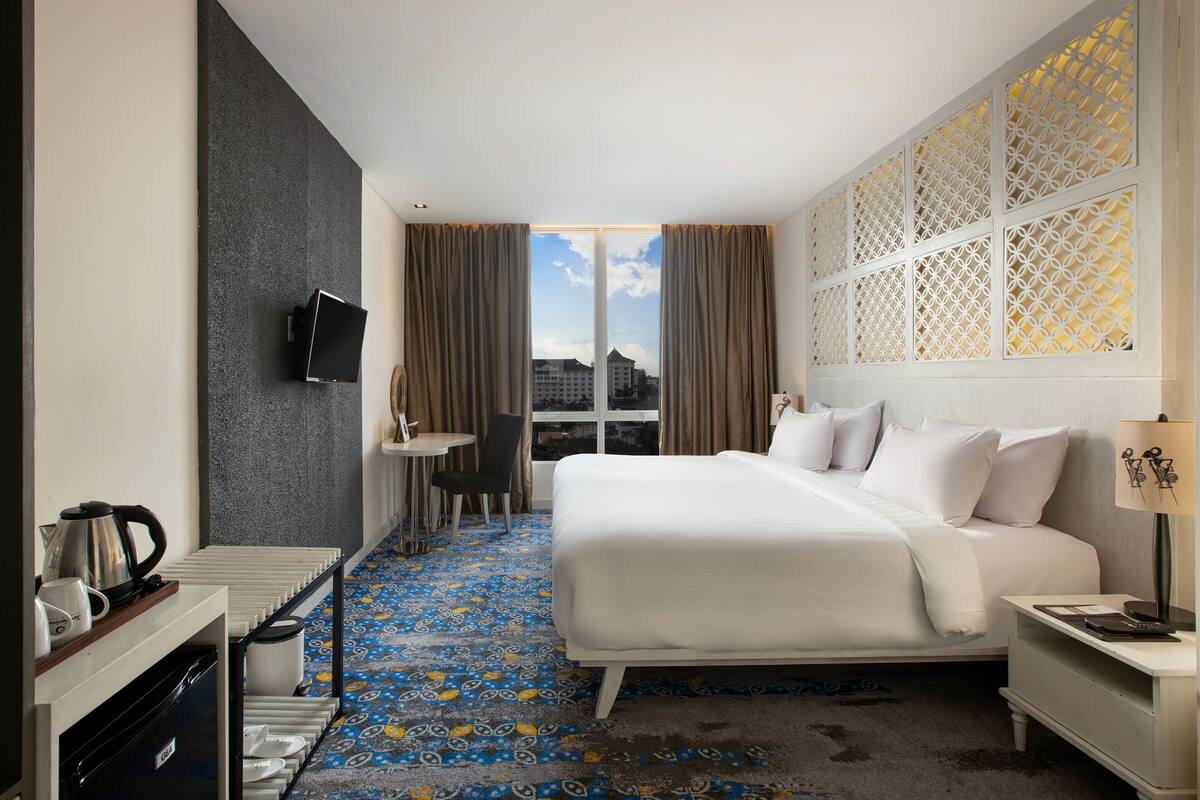 Une chambre d’hôtel comprenant un lit, un bureau avec une chaise, un téléviseur fixé au mur et une fenêtre avec des rideaux.