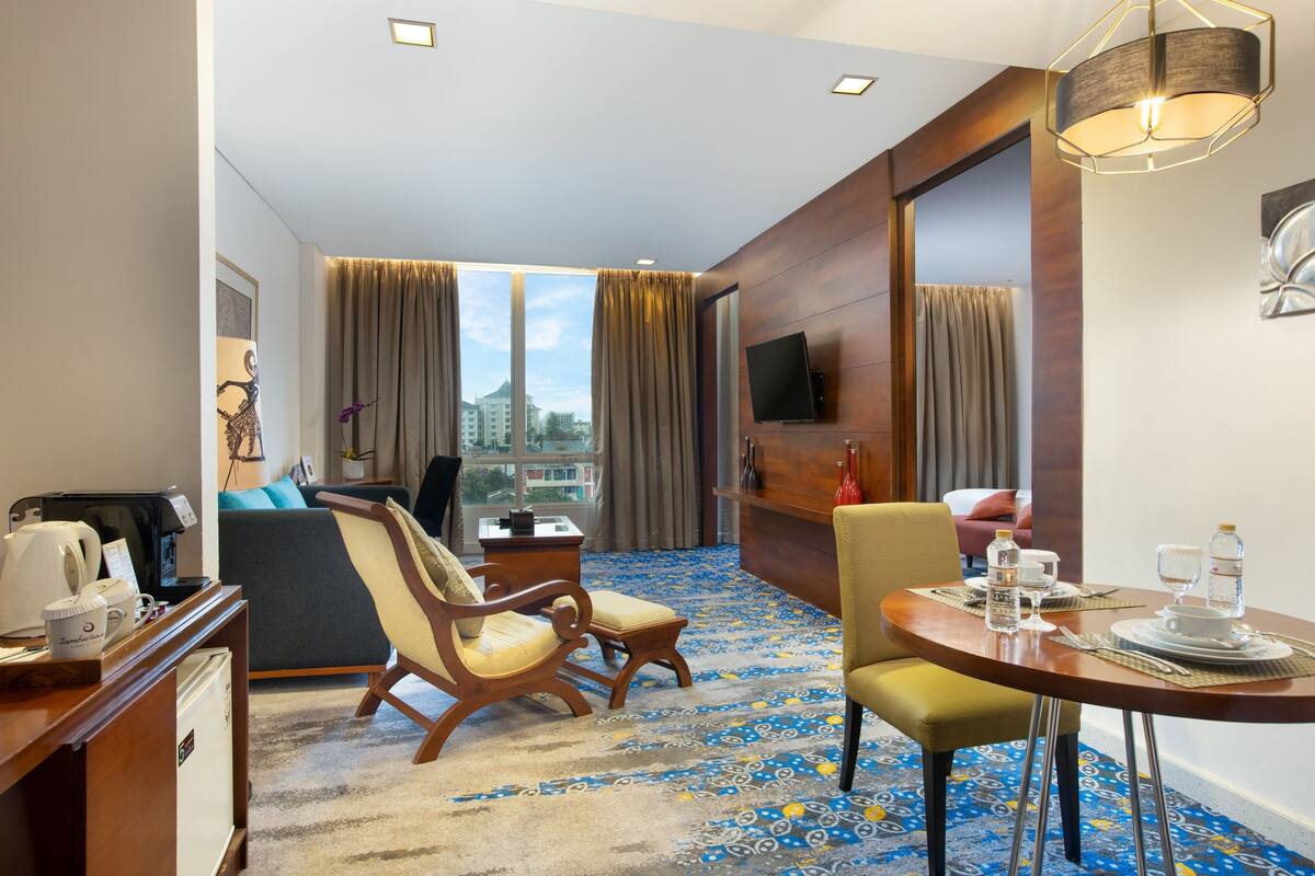 Une chambre d’hôtel dotée d’une grande fenêtre, d’une table à manger dressée avec des verres et une bouteille d’eau, d’un canapé et d’un bureau avec une machine à café.