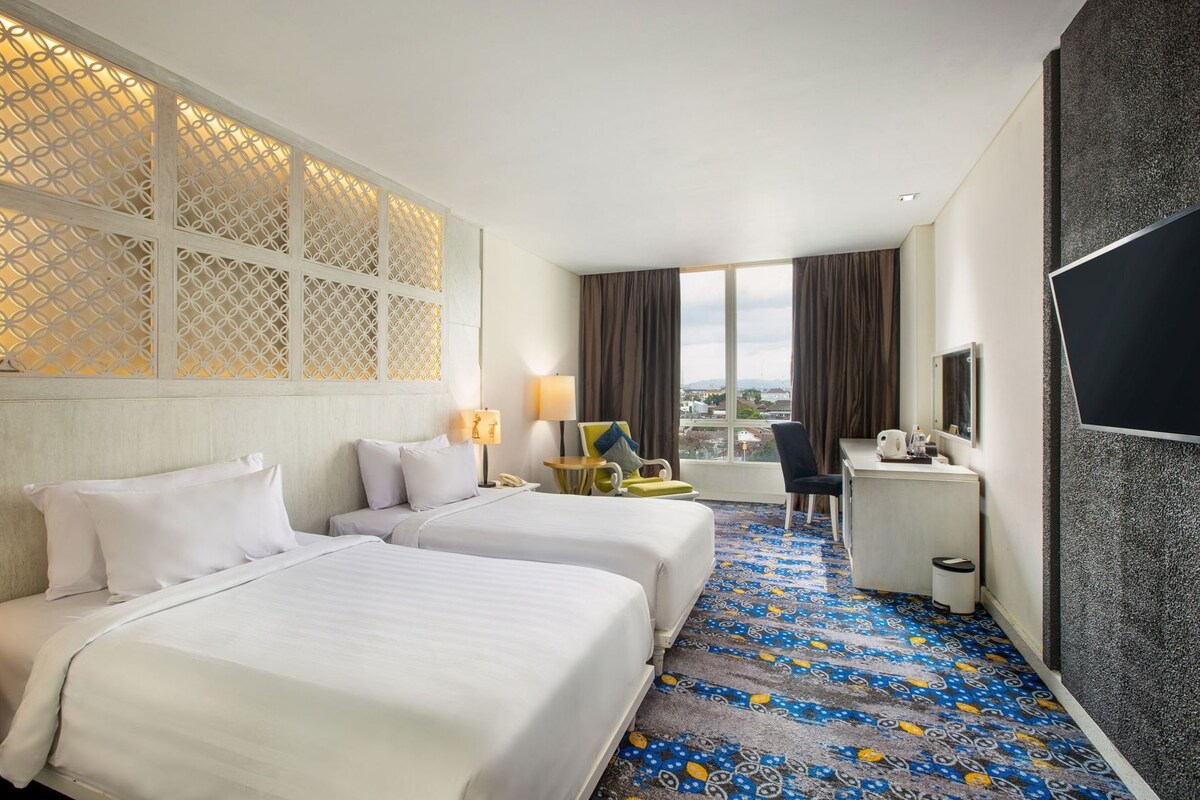 Une chambre d’hôtel avec deux lits, un bureau, une chaise, une télévision et une grande fenêtre avec des rideaux.