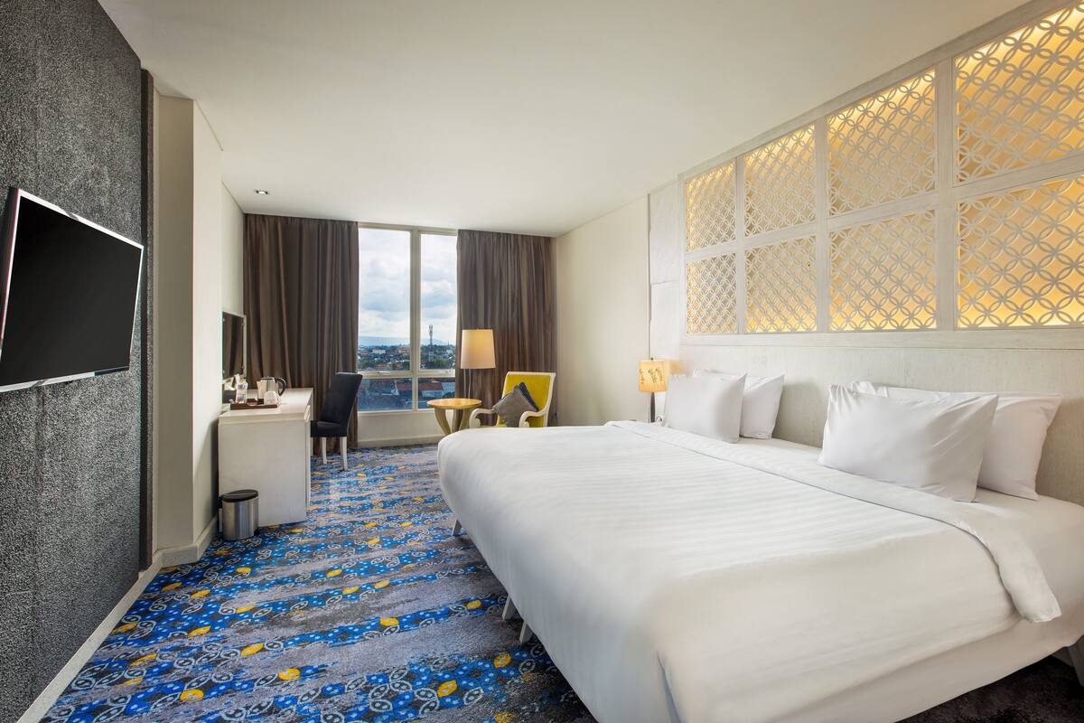 Une chambre d’hôtel avec un grand lit, un bureau, une chaise, une télévision et une vue sur la ville grâce à de grandes fenêtres.