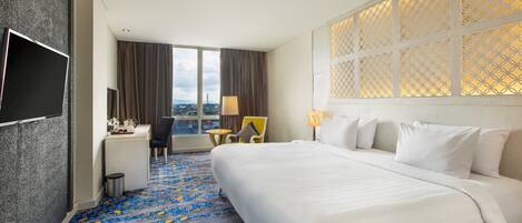 A hotel room with a large bed, a desk, a chair, a TV, and a view of the city through large windows.