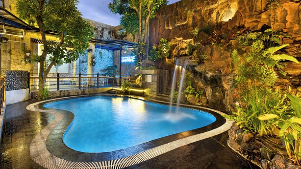 An indoor swimming pool with a waterfall feature, surrounded by tropical plants and a rocky wall.