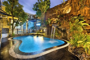 An indoor swimming pool with a waterfall feature, surrounded by tropical plants and a rocky wall.