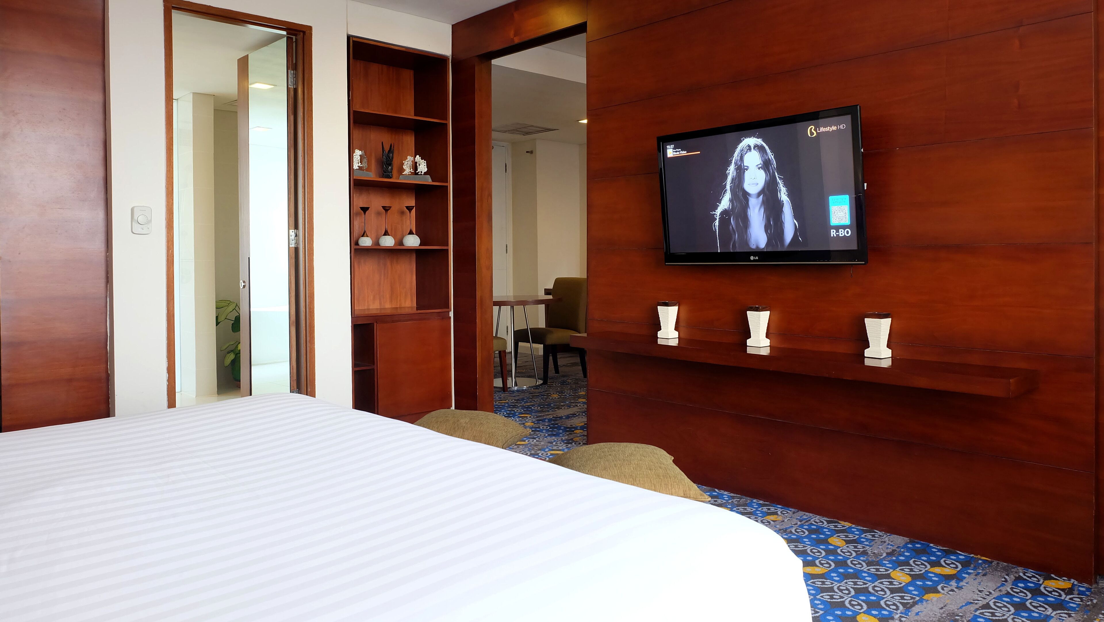 A hotel room with a bed, a TV mounted on the wall, a wooden wardrobe, and a small table with chairs.