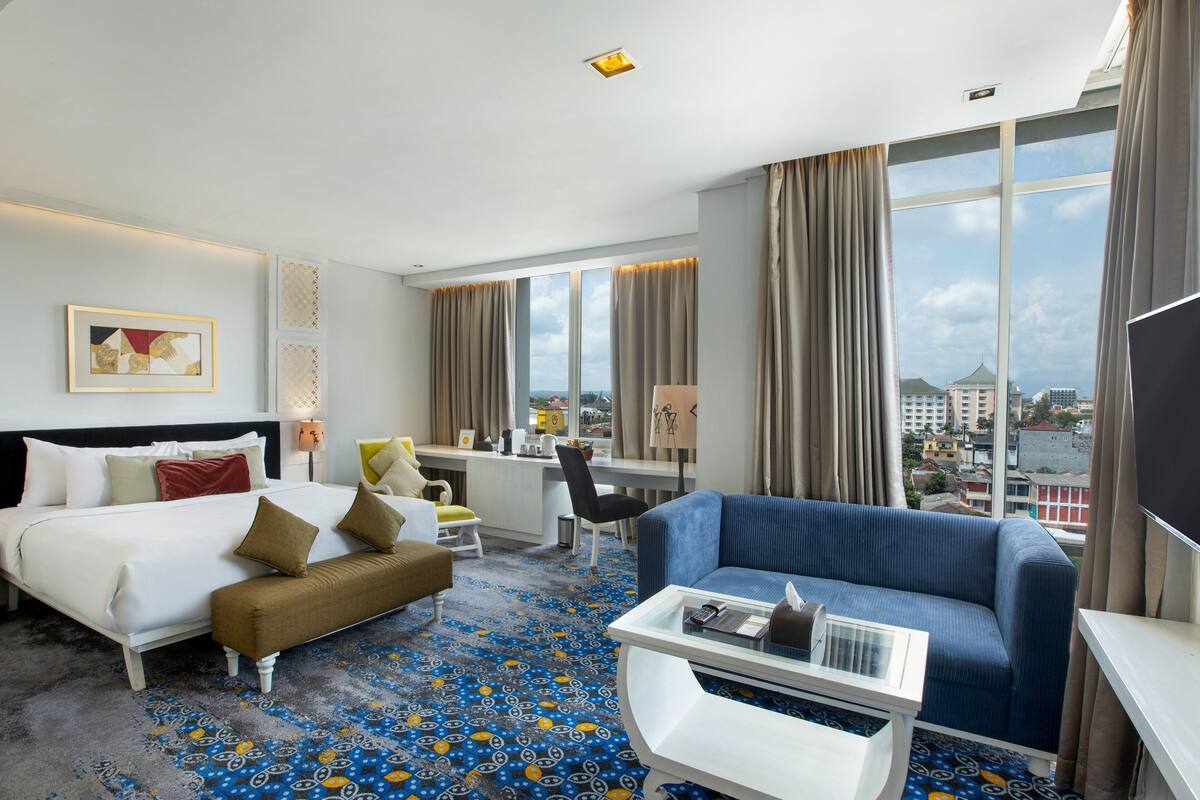 Une chambre d’hôtel équipée d’un lit, d’un canapé, d’un bureau et d’une grande fenêtre donnant sur la ville.