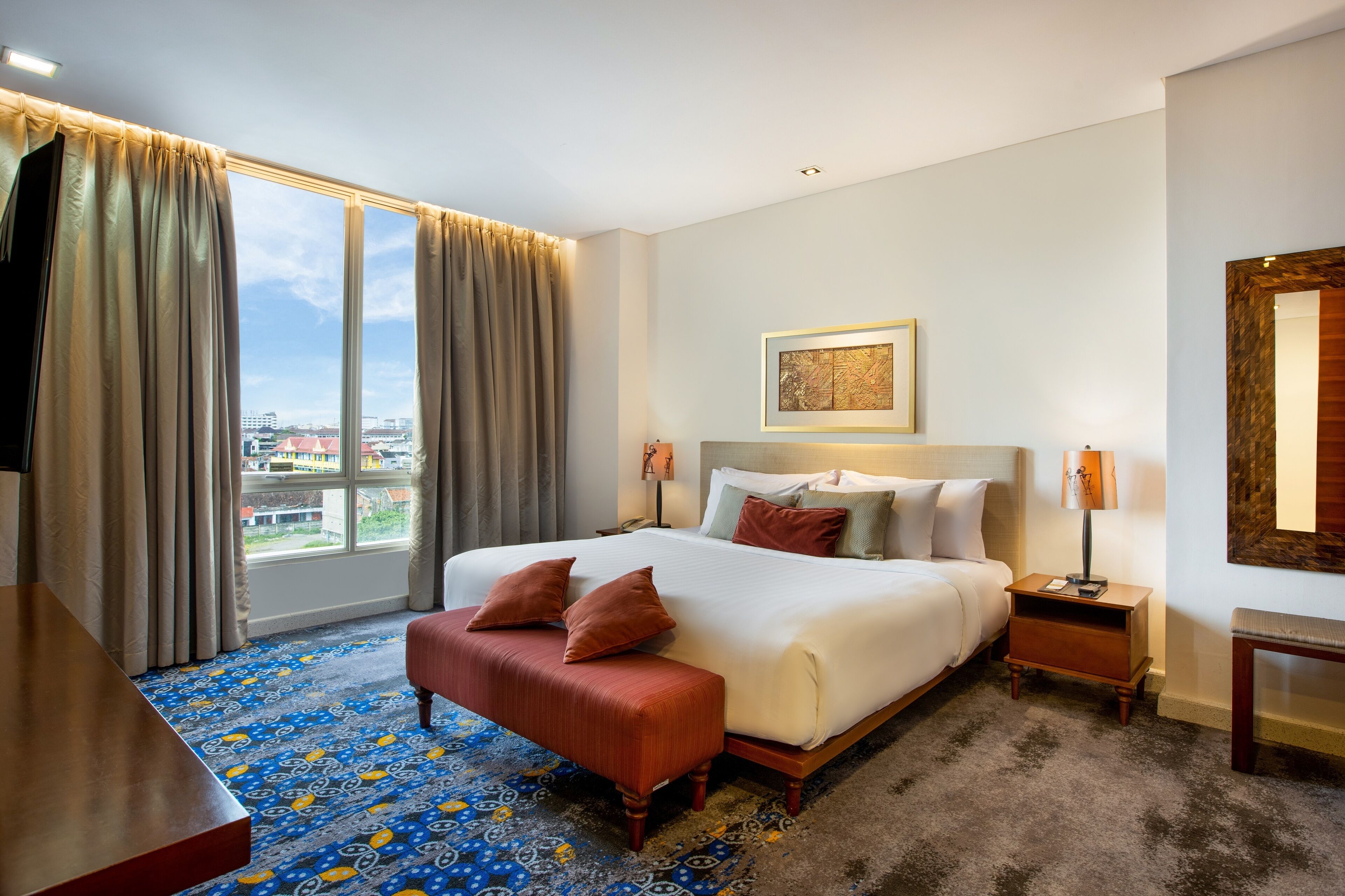 A hotel room with a large bed, a desk, a chair, a nightstand, a lamp, and a view of the city through the window.