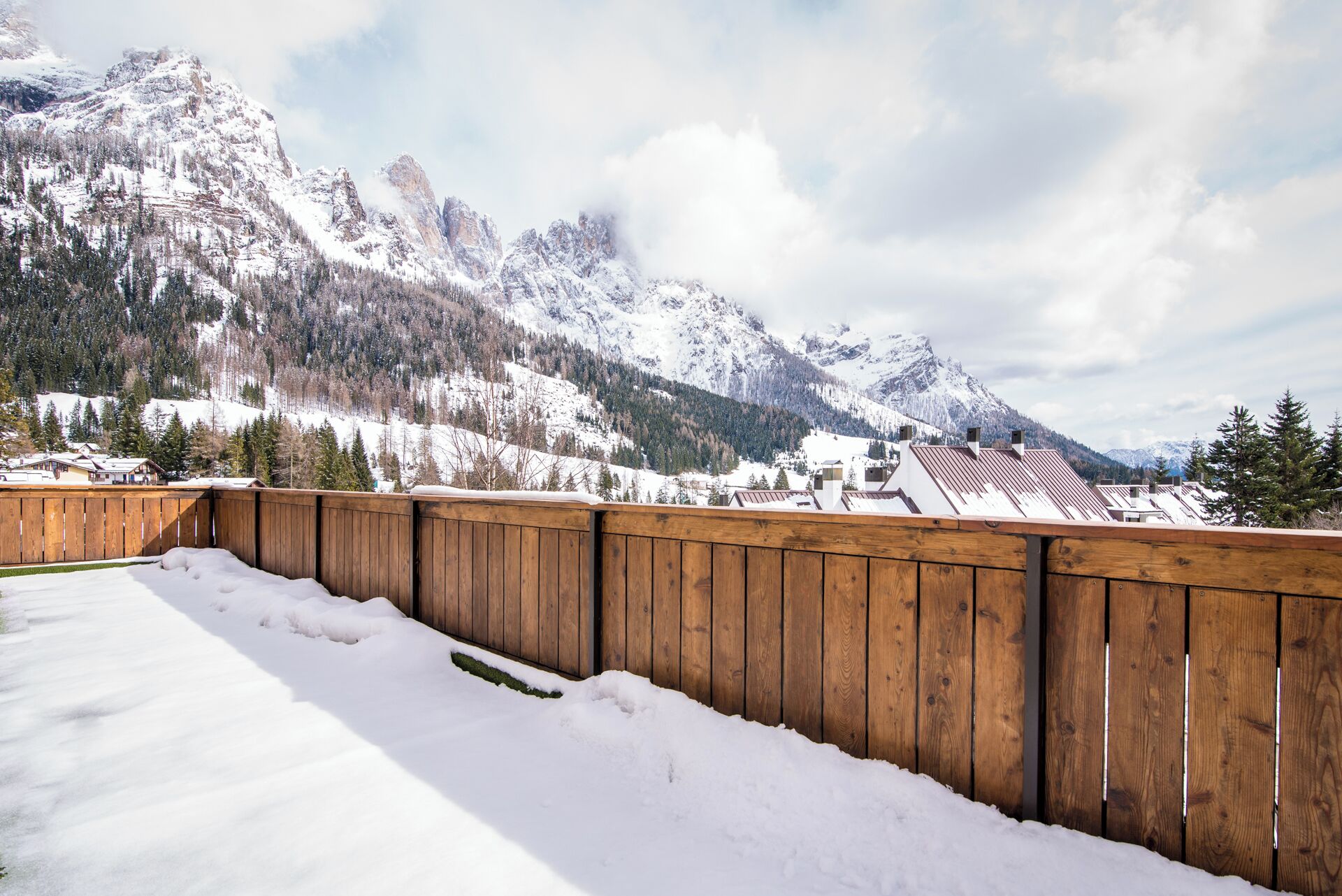 A wooden deck with snow-covered ground, overlooking a mountainous landscape with snow-covered peaks and a clear sky.
