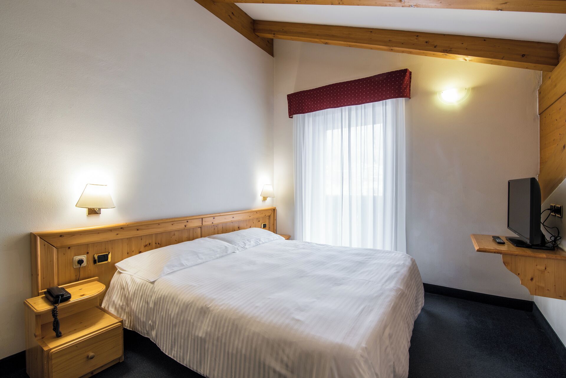 A hotel room with a wooden headboard, a bed with white bedding, a bedside table with a lamp, a TV on a wooden stand, and a window with sheer curtains.