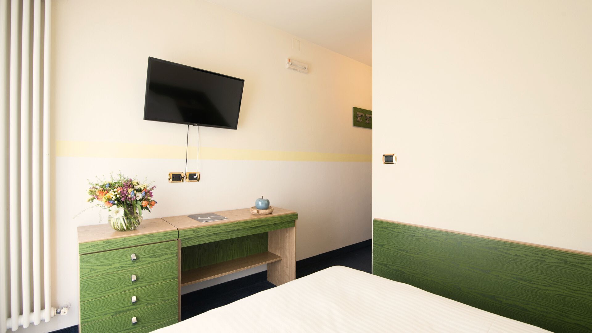 Een hotelkamer met een bed, een bureau met lades, een flatscreen televisie aan de muur en een vaas met bloemen op het bureau.