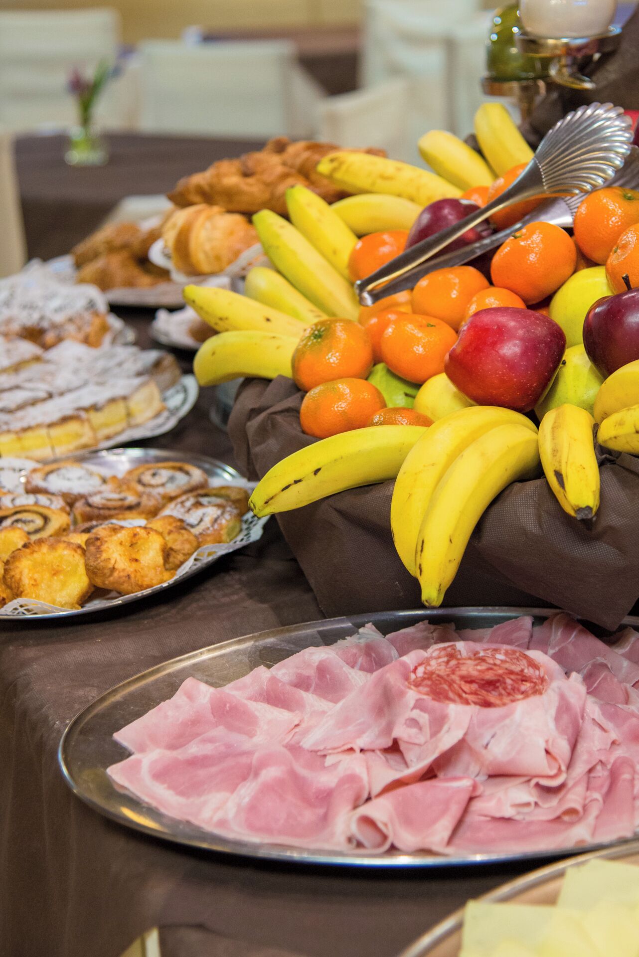 A buffet table with various food items including bananas, oranges, apples, pastries, and sliced meats.