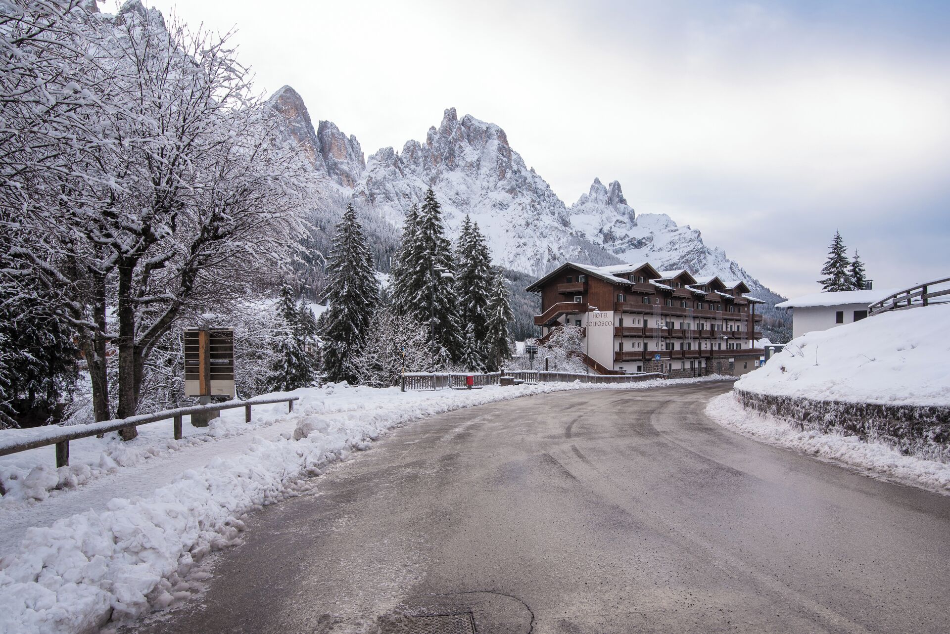 Une route de montagne enneigée menant à un hôtel à étages et doté de balcons, entouré d’arbres et de montagnes couverts de neige.