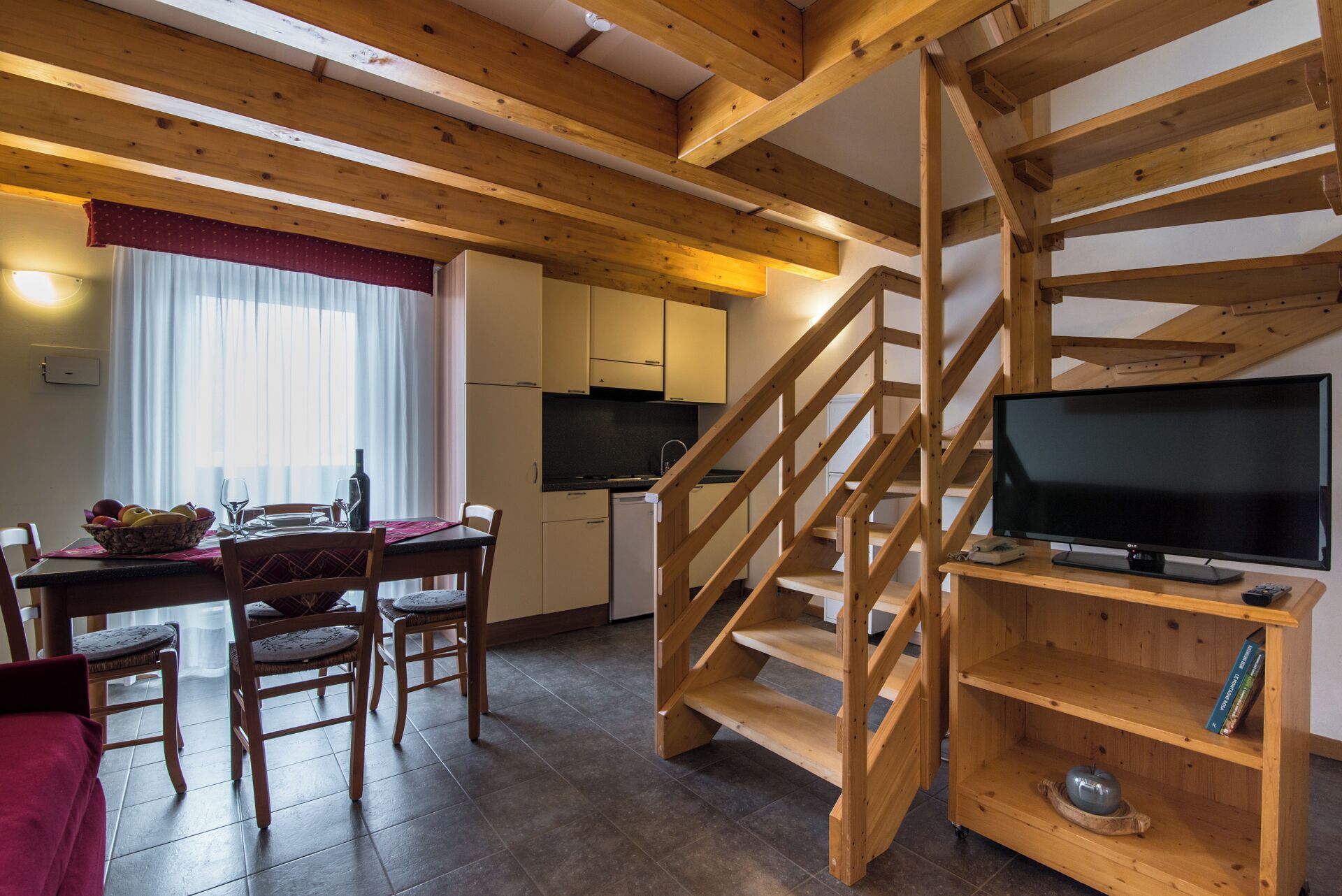 A compact living space with a wooden staircase, a dining area with chairs and a table, a kitchen with cabinets, and a TV on a wooden stand.