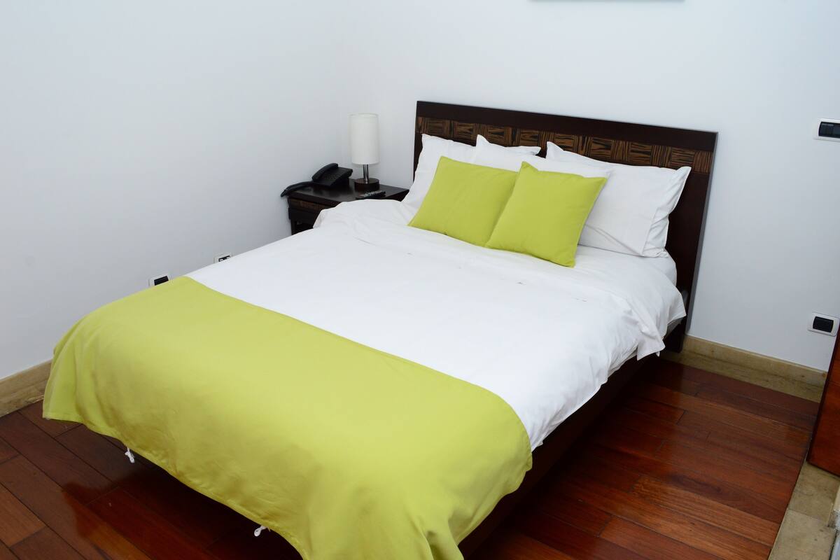 Un lit bien fait, recouvert d’une couvre-lit blanche et verte, avec une tête de lit en bois et une table de chevet sur laquelle se trouvent une lampe et un téléphone.