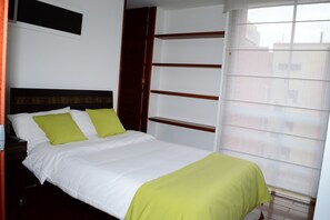 A neatly made bed with white and green bedding, a wooden headboard, and a window with blinds.