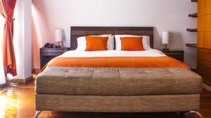 Una cama bien hecha con ropa de cama blanca y naranja, un cabecero de madera y un banco tapizado de color gris.