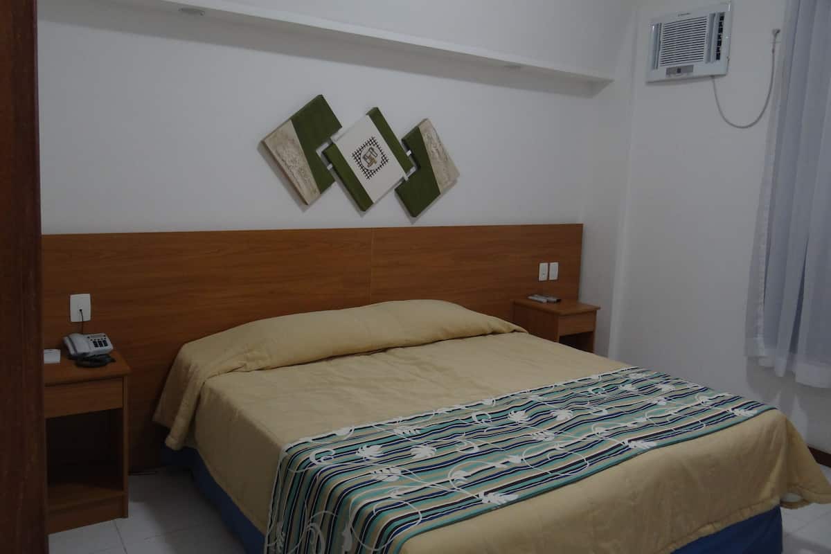 A hotel room with a wooden headboard, a bed with a beige bedspread, a patterned throw blanket, a bedside table with a phone, and a wall-mounted air conditioner.