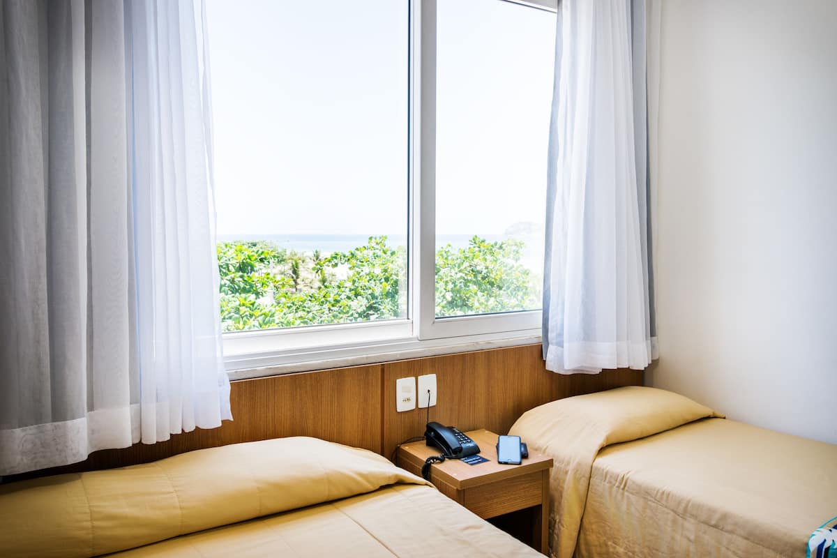 A hotel room with two beds, a wooden nightstand, a telephone, and a view of greenery and water outside the window.