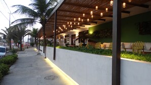 A covered outdoor seating area with hanging lights, palm trees, and a green wall with seating.