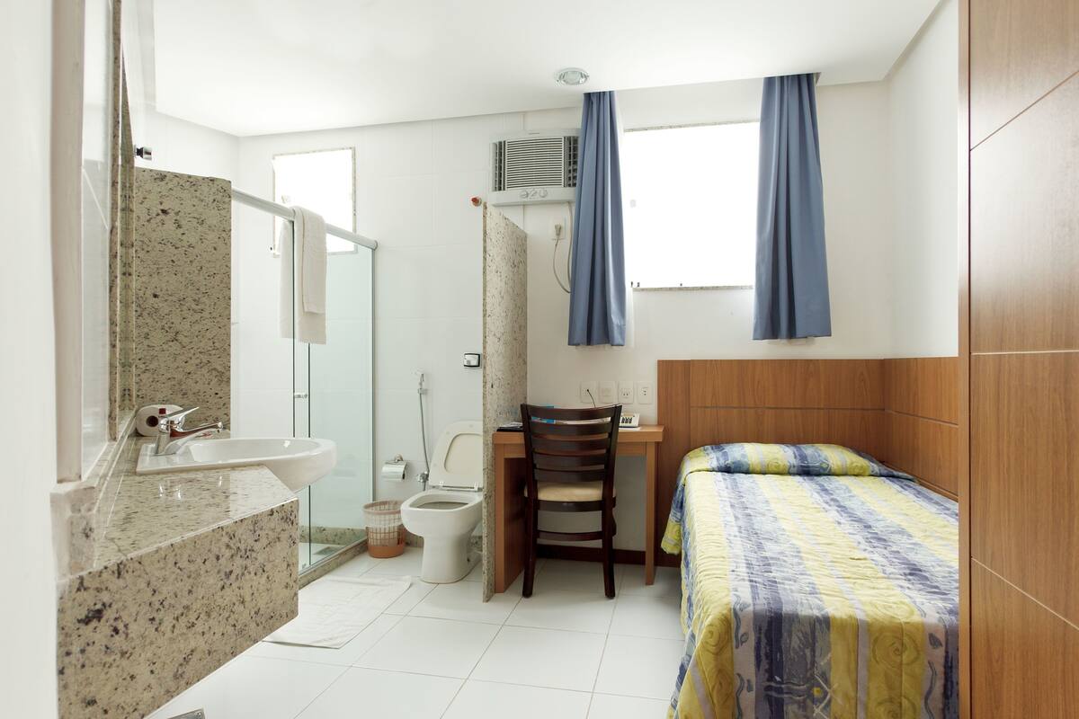 A bedroom with a bed, a desk, a chair, a bathroom with a sink, and a shower.