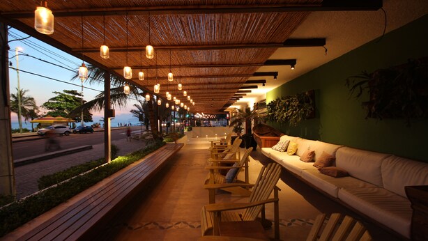 A covered outdoor seating area with wooden chairs and a white sofa, overlooking a street with palm trees and cars. - Praia Linda (Rio de Janeiro)