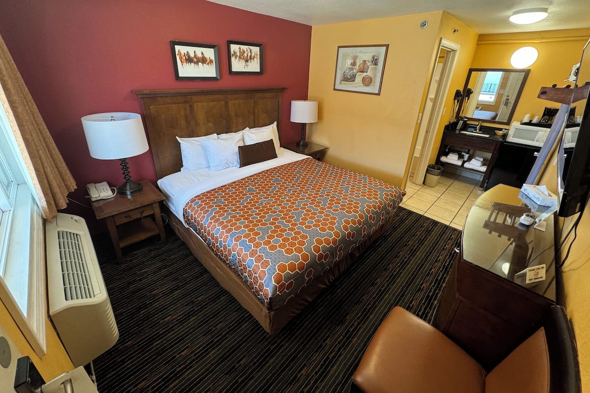 A hotel room with a large bed, a wooden headboard, a desk with a lamp, a telephone, and a mirror. There are pictures on the wall and a bathroom visible through an open door.
