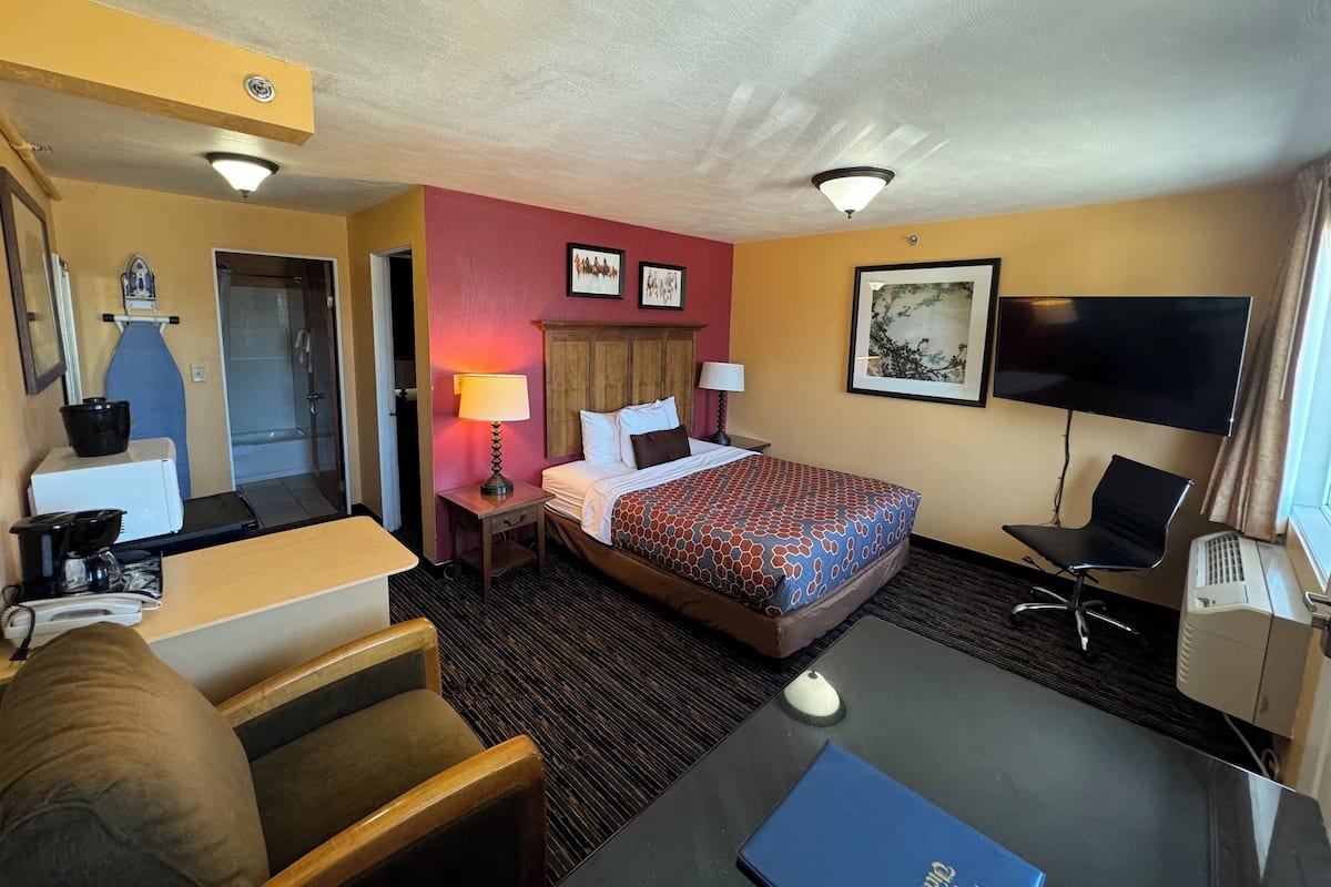 A hotel room with a bed, desk, chair, TV, and a small kitchenette.