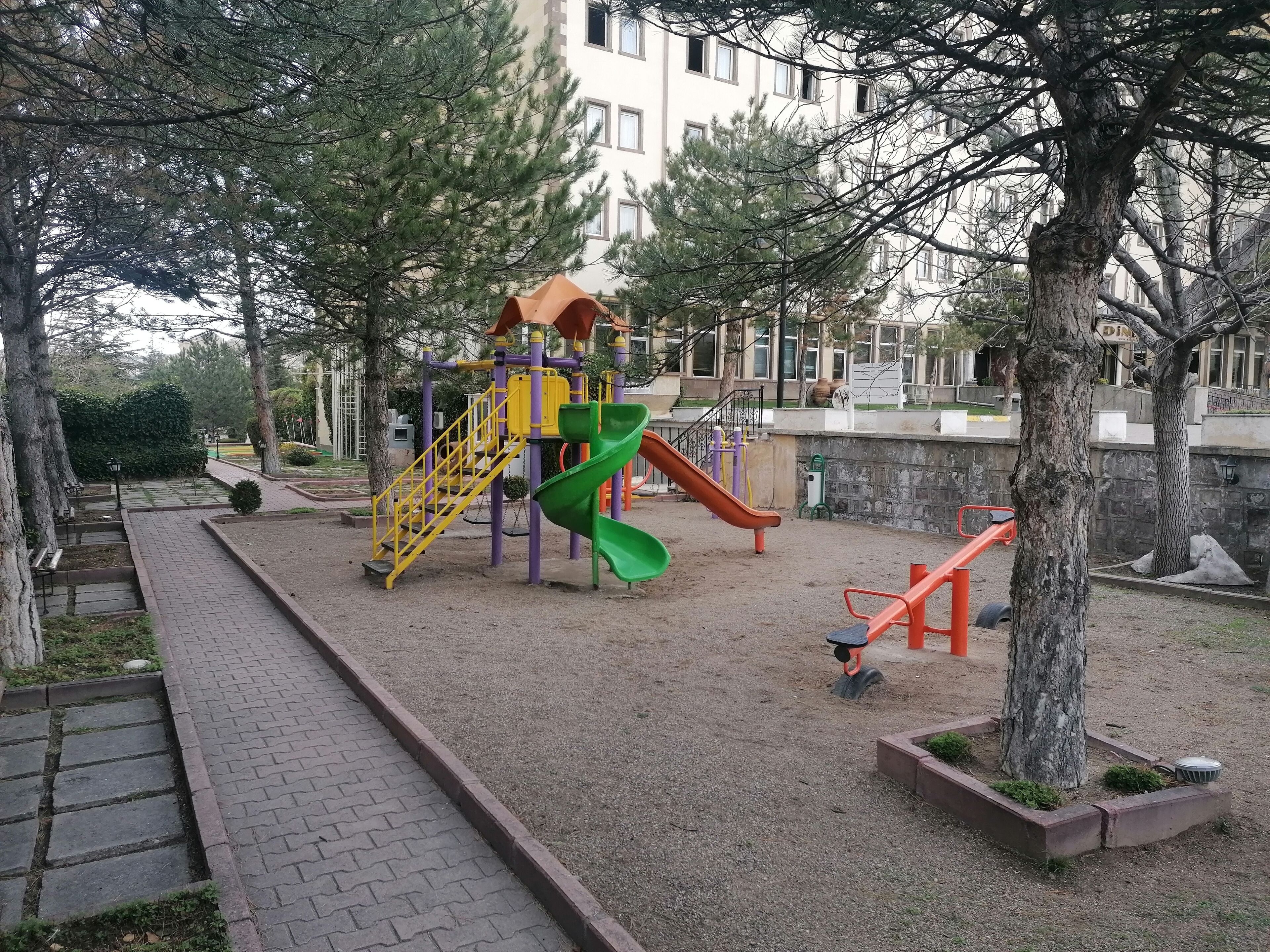 A playground with a slide and climbing structures, surrounded by trees and a paved walkway.