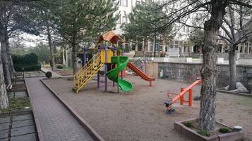 A playground with a slide and climbing structures, surrounded by trees and a paved walkway.