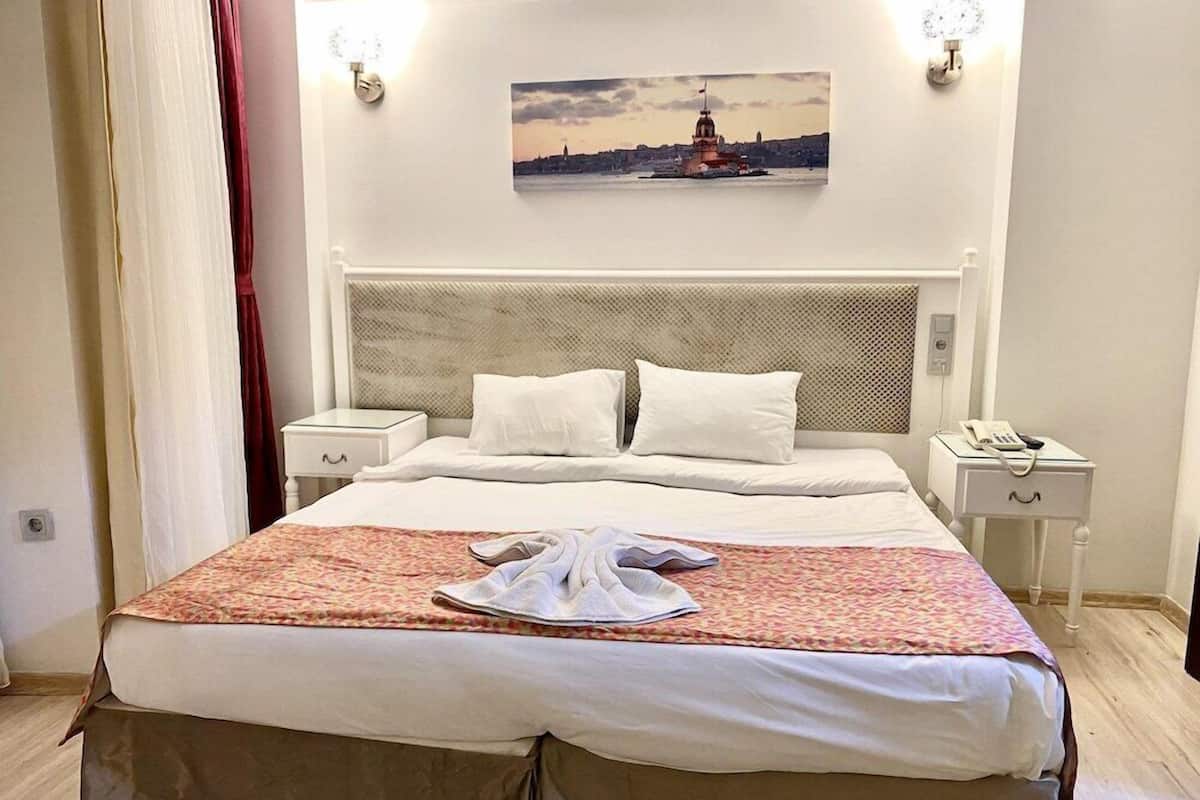 Habitación de hotel con cama, mesita de noche, lámpara, un cuadro en la pared y ventana con cortinas.