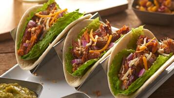 Three tacos with lettuce, red onions, and shredded cheese on a white tray, accompanied by a glass of iced beverage and lime wedges.
