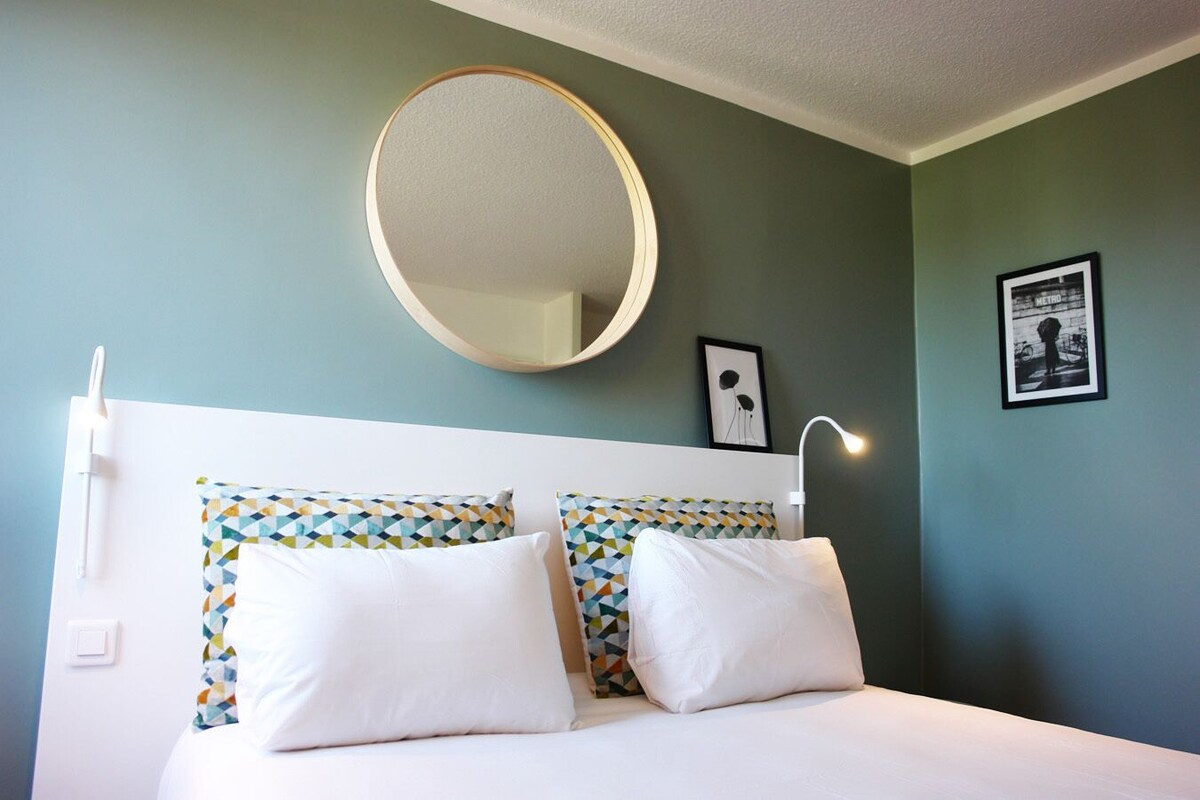 A bedroom with a bed, two pillows, a round mirror, a wall-mounted light, and a framed picture.