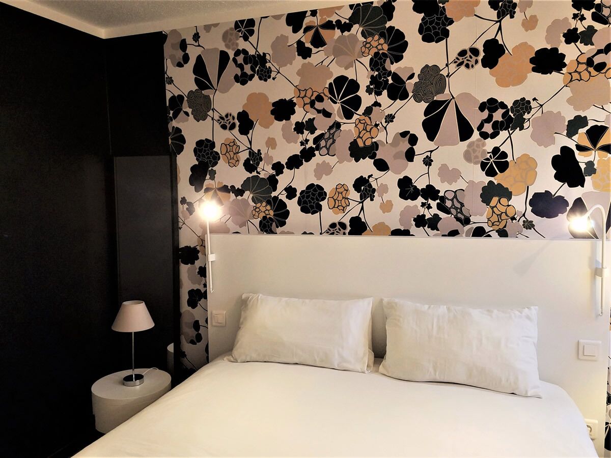 A bedroom with a bed, two pillows, a nightstand with a lamp, and a wall with a floral pattern.