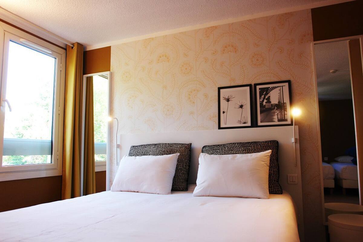 Ein Hotelzimmer mit einem großen Bett, zwei Kissen, einer Kopfteil, einem Fenster mit Vorhängen und einer Wand mit floralem Tapetenmuster sowie zwei gerahmten Bildern.
