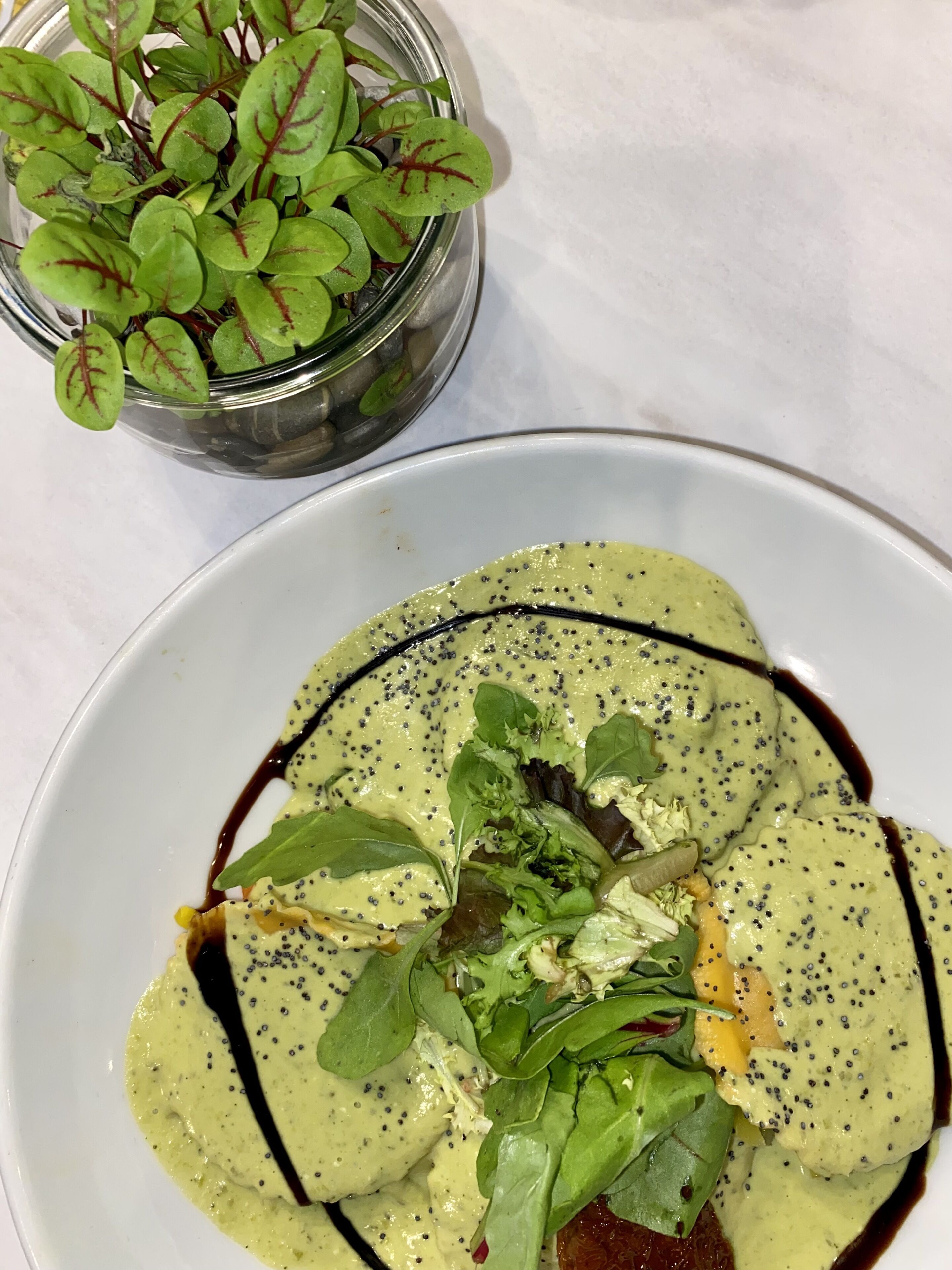 Un plato de comida con salsa verde, vegetales de hoja y un frasco pequeño de vegetales al fondo.