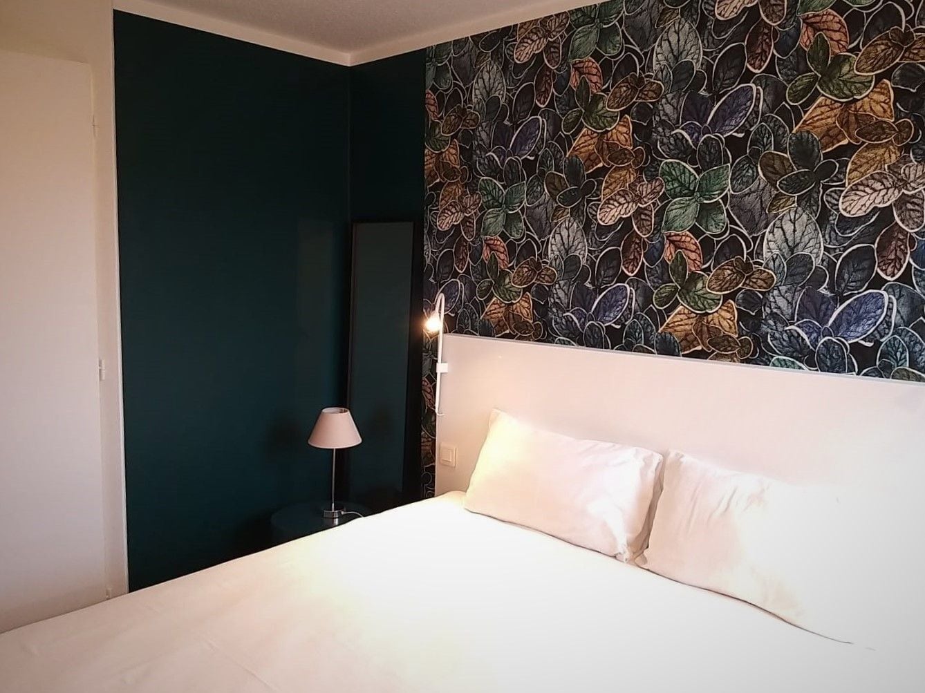 A hotel room with a bed, a nightstand, a lamp, and a wall with a leaf pattern.