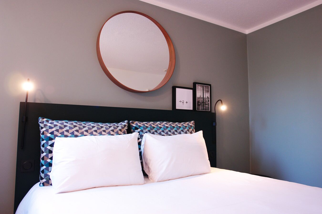 A bed with white bedding, a geometric patterned pillow, a round mirror, and two wall-mounted lights.