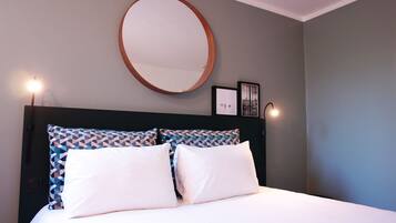 A bed with white bedding, a geometric patterned pillow, a round mirror, and two wall-mounted lights.