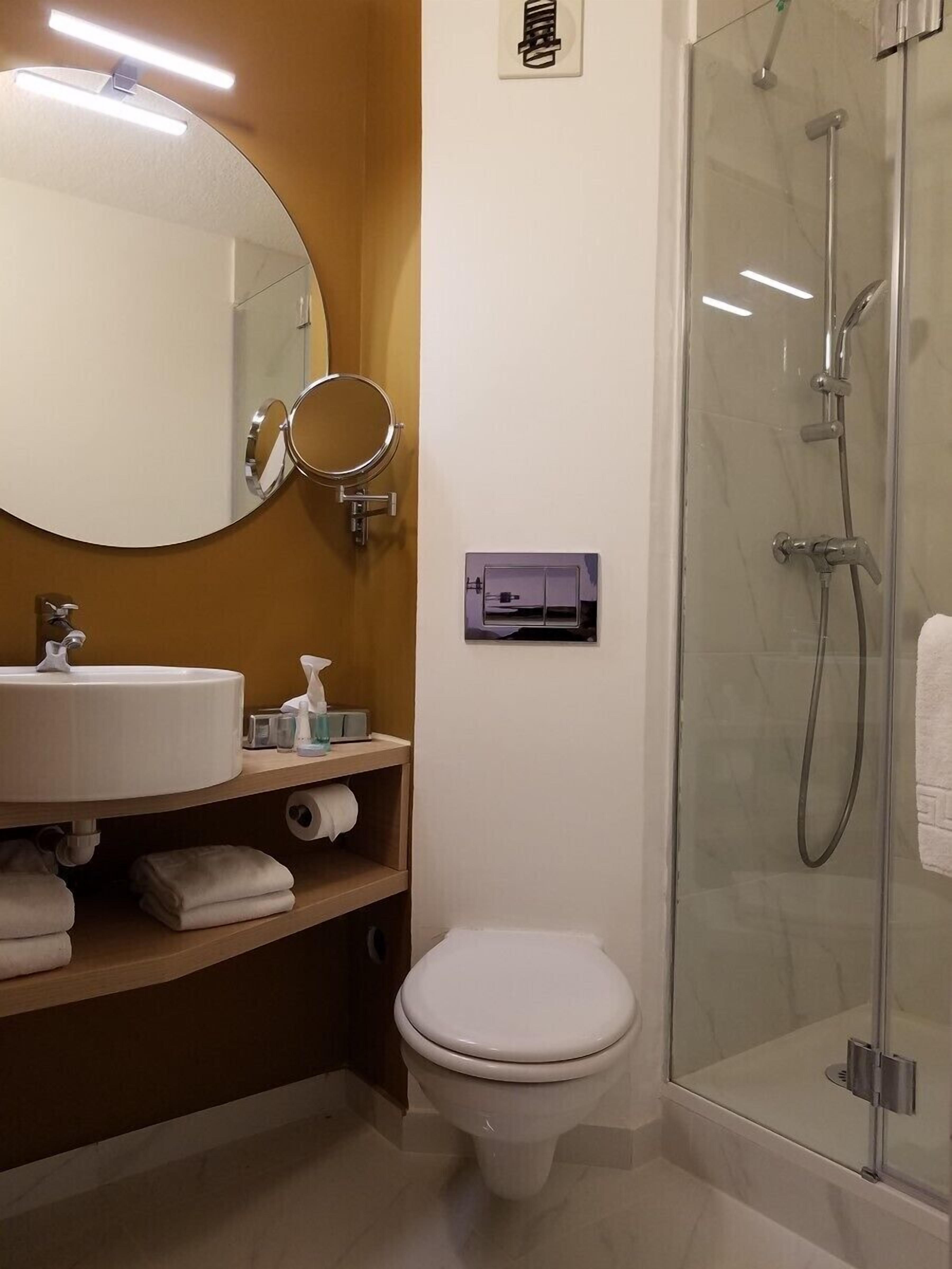 Un baño con espejo redondo, lavamanos, inodoro y cabina de ducha con mampara de vidrio.