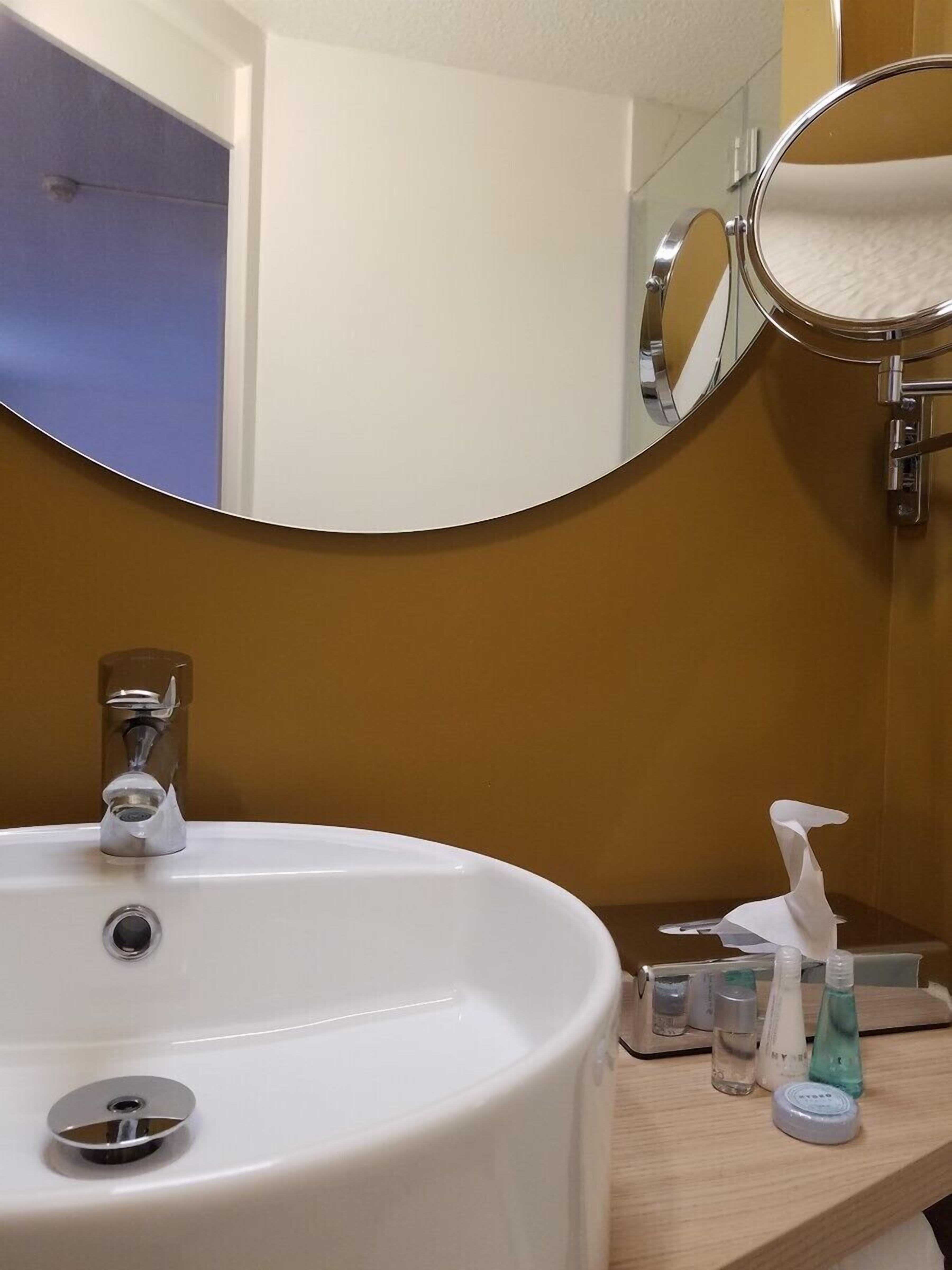 Kylpyhuoneessa on valkoinen pesuallas, peili ja hygieniatuotteita tasolla.