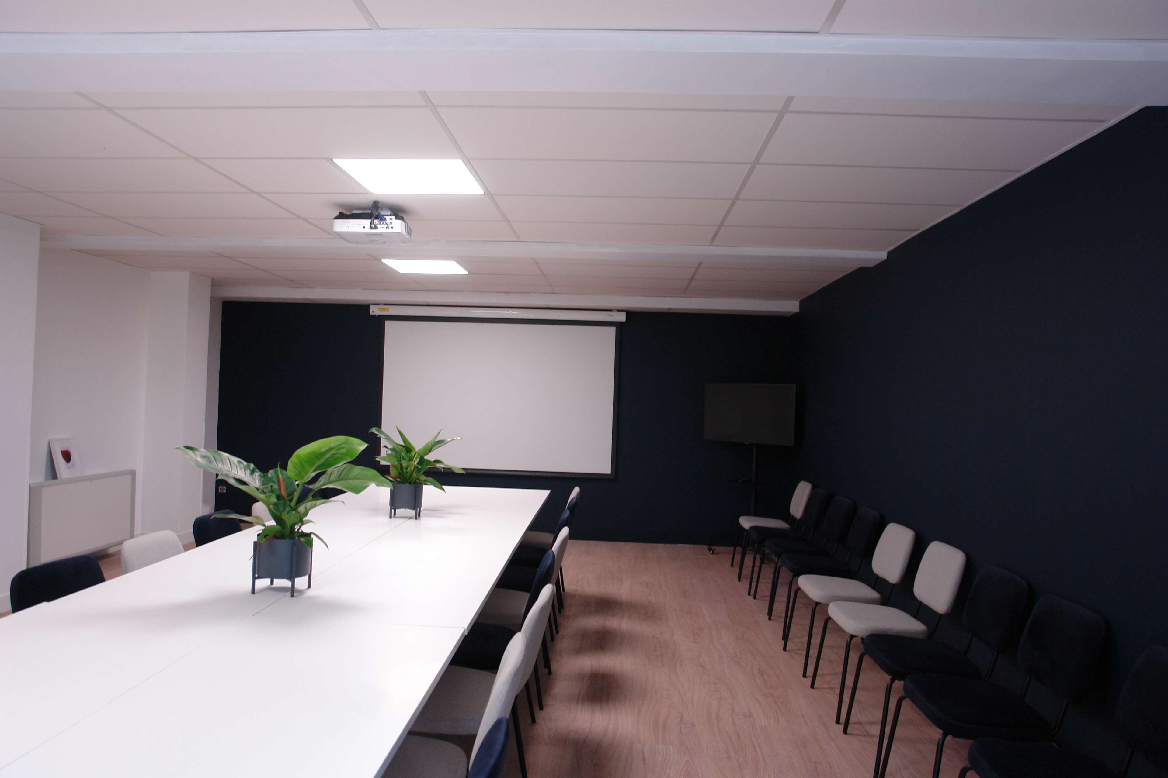 Un salón de conferencias con una larga mesa, sillas, un proyector y una pantalla.