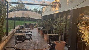 Um pátio ao ar livre com mesas redondas de madeira e cadeiras pretas, rodeado por plantas em vasos e uma pérgola com toldo branco.