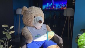 Um urso de pelúcia grande sentado numa cadeira, vestindo uma camisa 'BRIT HOTEL', cercado por plantas de interior e uma vista urbana ao fundo.