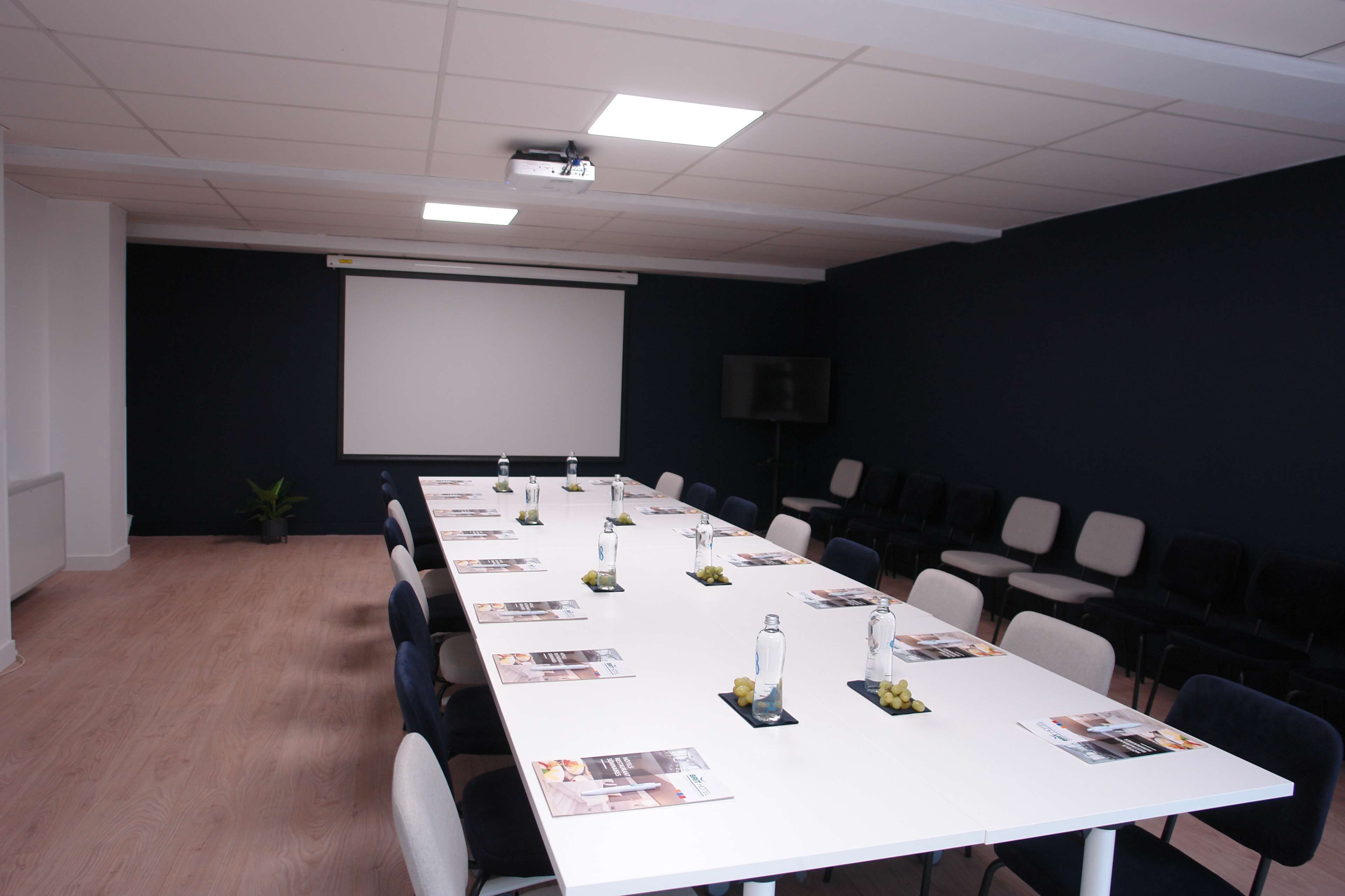 Un salón de conferencias con una larga mesa, sillas, una pantalla de proyector y una televisión.