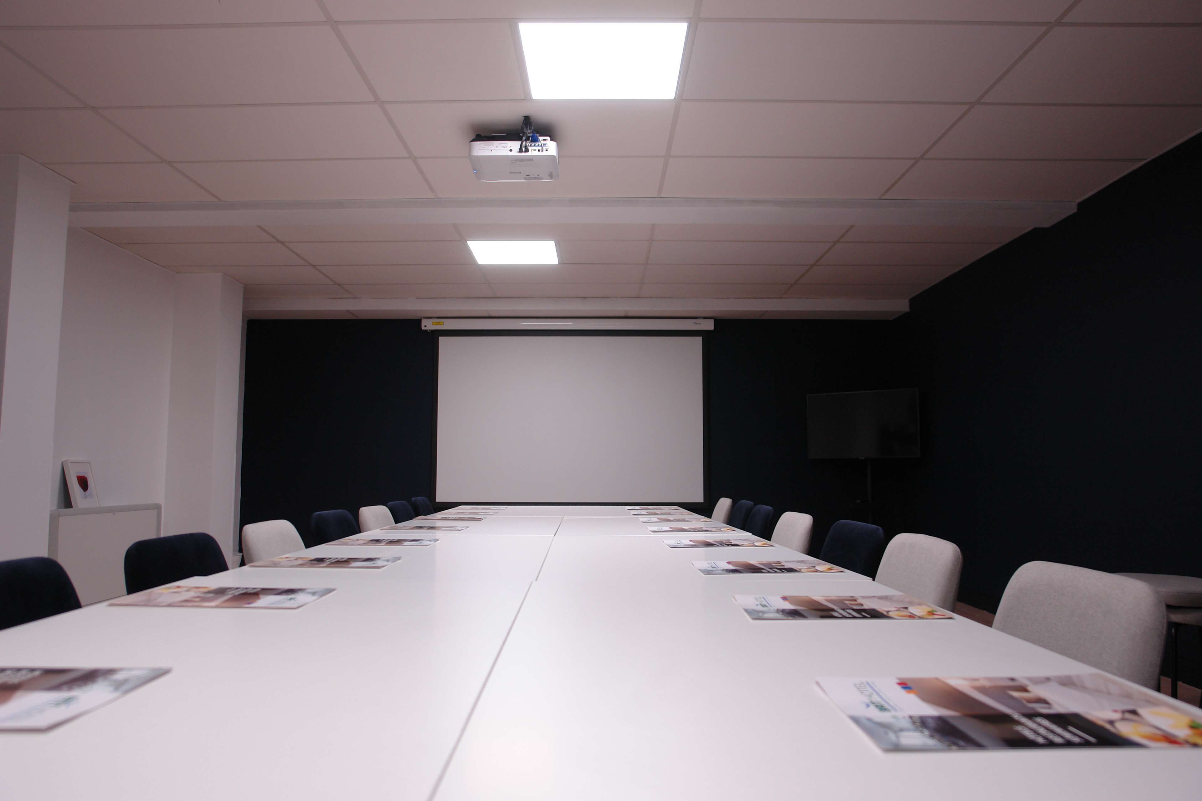 Een vergaderzaal met een lange tafel, stoelen, een projecter en een scherm.